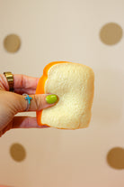 Close-up of a hand pressing a toast-shaped squishy with a cream-colored center and golden-orange crust.
