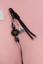 Black heart keychain charm with checkered beads, mini handbag charm and knotted cords on a pink background.