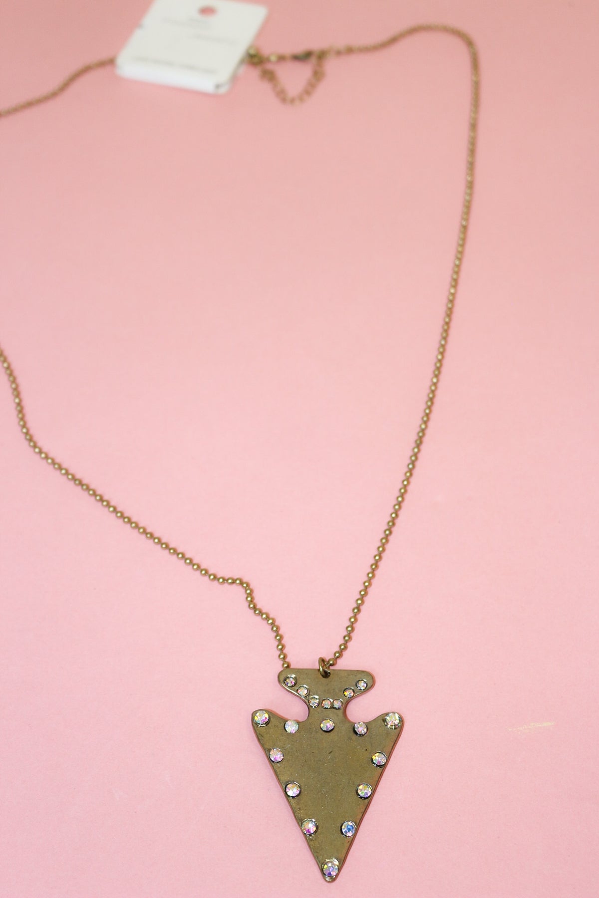 Gold-tone arrowhead necklace with iridescent jewel accents, fine-link chain, and extender displayed on a pink background.