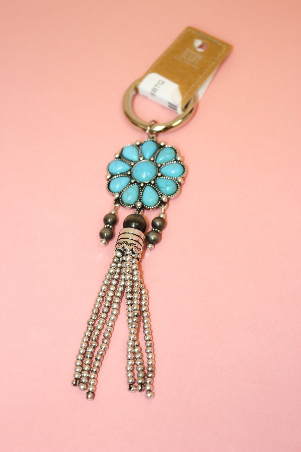 Turquoise-colored flower keychain with dark bead accents, silver-tone beaded tassel, and round key ring on pink background.