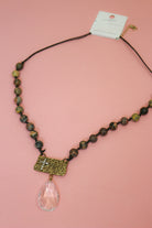 Brown and earthy mixed-tone beaded necklace on dark cord with faith plaque and clear teardrop pendant on pink background.