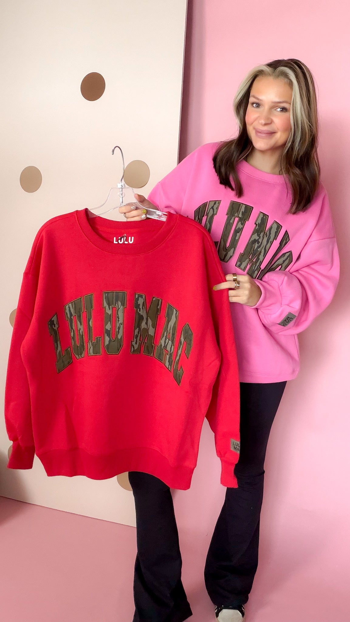 Model wearing a pink Lulu Mac camo-letter sweatshirt while holding the red version on a hanger in front of her.