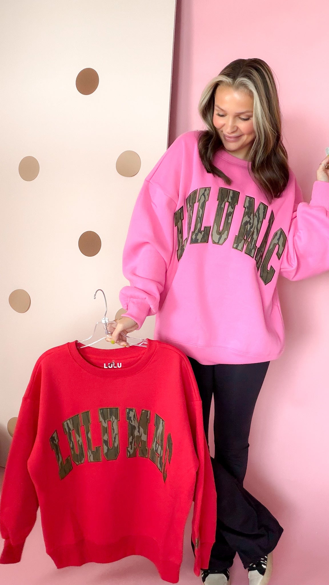 Model in a pink Lulu Mac sweatshirt and black pants holding the red Bottomland camo-letter sweatshirt beside her.