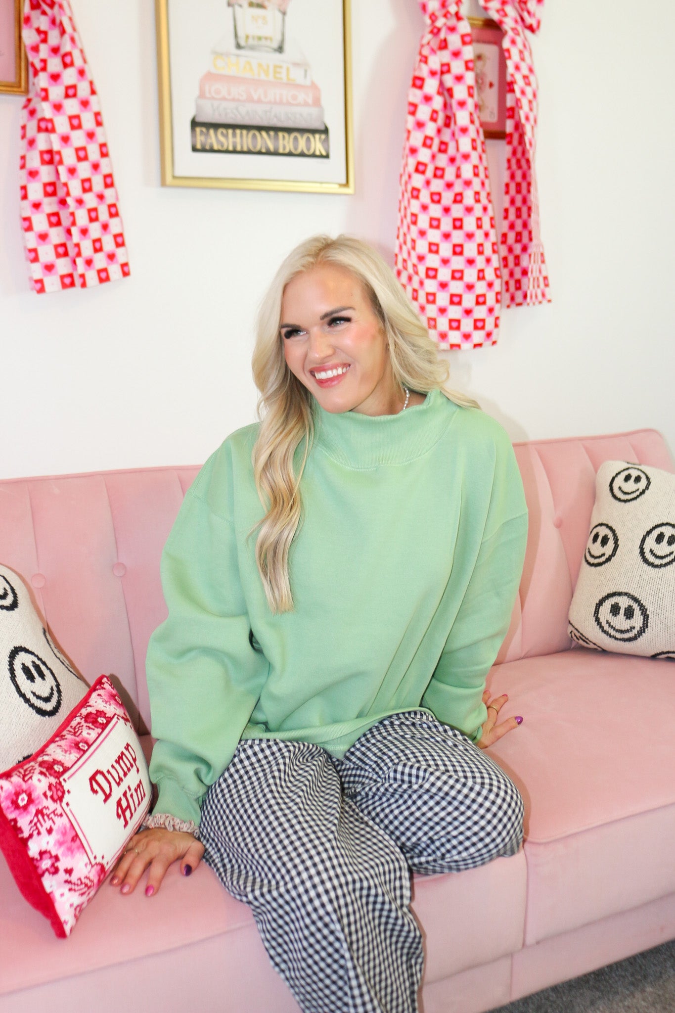 Woman in a green sweater and checkered pants standing in a room with pink couch, decorative pillows, and wall art.