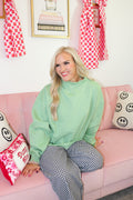 Woman in a green sweater and checkered pants standing in a room with pink couch, decorative pillows, and wall art.