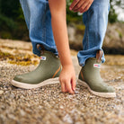 Child wearing olive XTRATUF Kids' Ankle Deck Boot Sport boots with jeans while exploring a rocky shore.