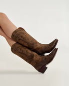Brown suede boots worn by a person on a light gray background. Tecovas women’s cowboy boots