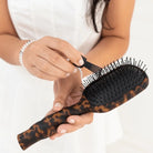 Hands using the cleaning tool on the Teleties tortoise Detangler Brush bristles